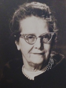 This black and white portrait of Elizabeth Benson shows a white woman with 1960's-style-horn-rimmed glasses and pearl necklace smiling at the camera. Her dark hair is pulled back atop her head and a broad collar encircles her shoulders.