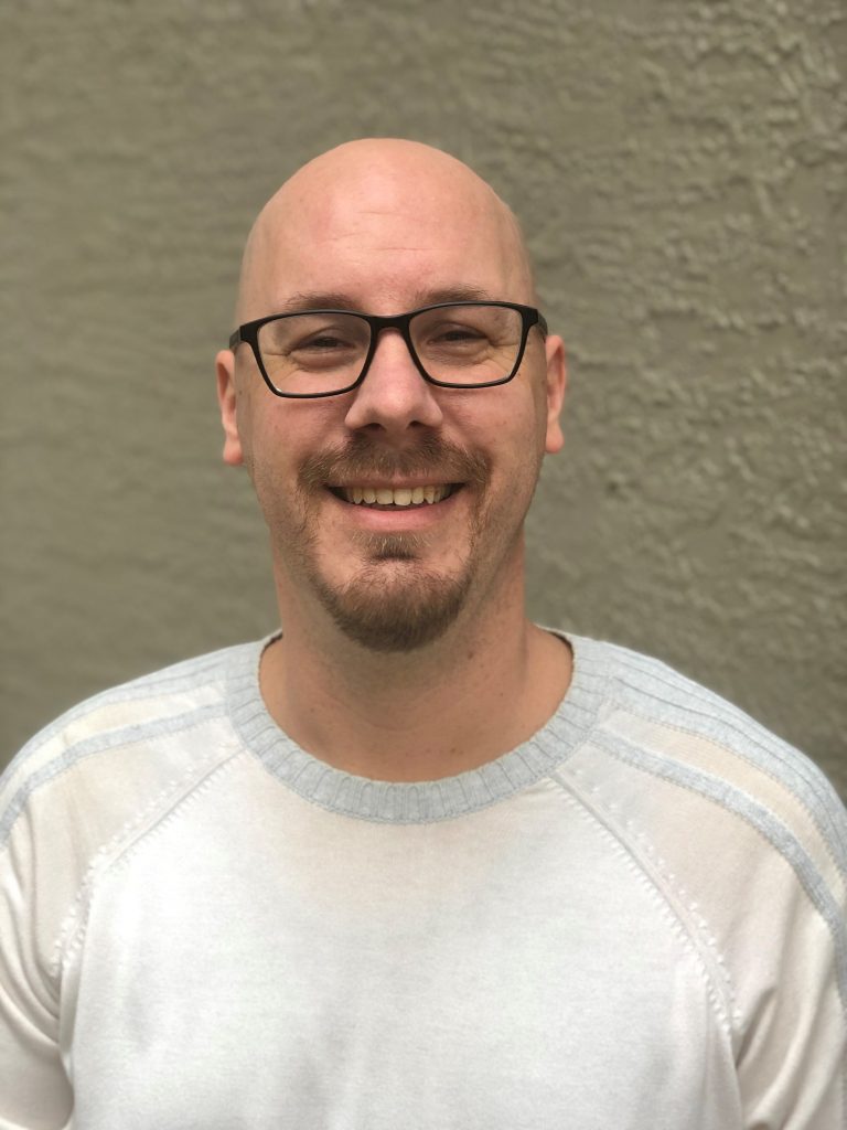 [Image description: A head shot of Vice President Robert Loeffler, smiling for a photo in front of a textured background. Bobby is a bald white male and is wearing a white and cream colored shirt with grey stripes on the shoulders with dark rimmed glasses.]