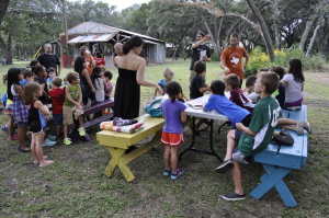 [Image description: Two separate tables are surrounded by children while a KODAheart member is signing directions. A volunteer is standing next to him wearing a burnt orange shirt.]