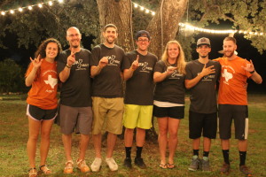 [Image description: KODAheart board members and volunteers stand side by side posing for a picture. On each end a KODAheart volunteer, dressed in burnt orange, is signing the sign for I-love-you. In between them the KODAheart board members dressed in gray, each fingerspelling one letter in the word Koda and a person signing the sign for heart. It is nighttime and the background is illuminated with white string lights.]