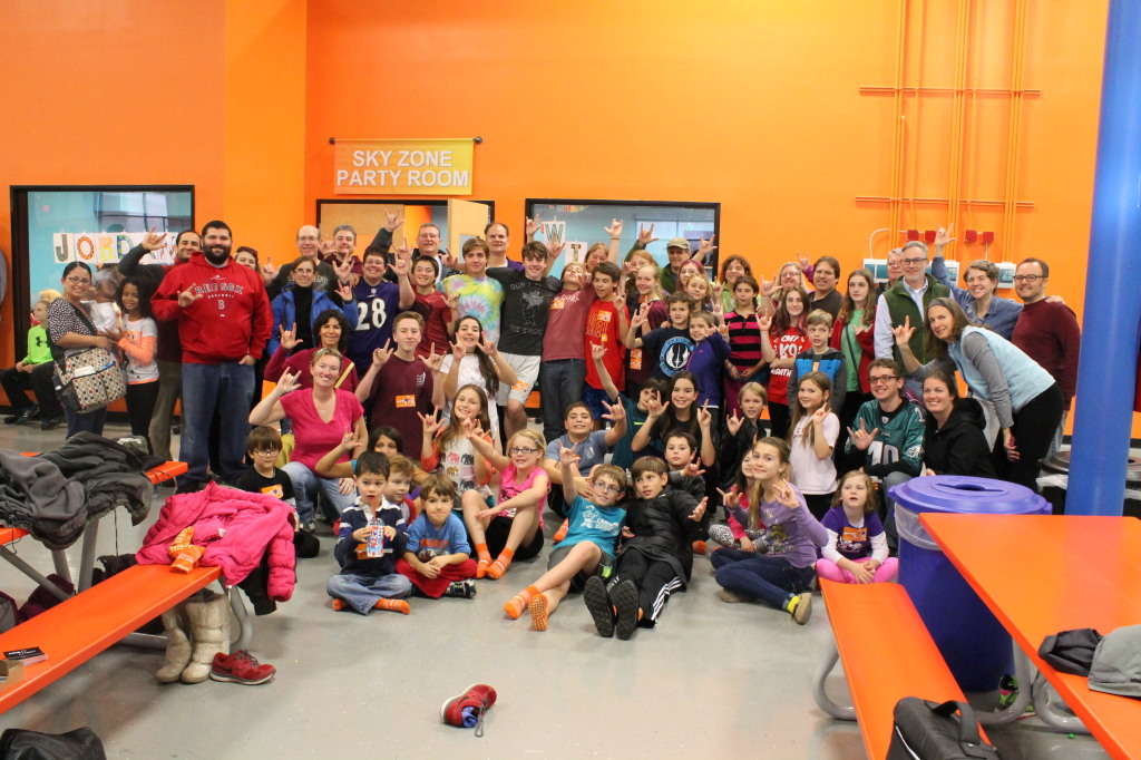 [Image description: A large num ber of f amilies are grouped together fo r a picture in front of a bright orange wall in SKY ZONE. An orange table can be seen in the foreground.]