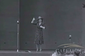 Image Description: A thin white woman in a polka dot dress stands on stage in front of a microphone. Her arms are moving as she signs.