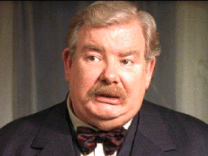 Image Description: A movie still of Richard Griffiths shows a white man with greying hair and a mustache wearing a suit jacket and bow tie. He is looking to the left with a concerned expression on his face.