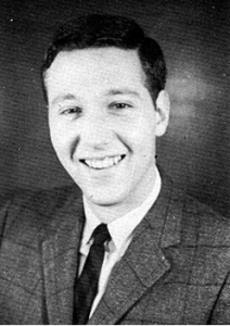 A young white man in a plaid suit jacket and dark tie, smiles pleasantly at the camera.