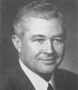 A black and white image of a white man with a suit and tie, smiling at the camera.