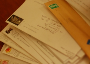 [Image Description: Several letters are placed on top of each other in a pile with stamps and mailing addresses. They are all white envelopes but one is a large orange mailing envelope.]