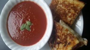 Image Description: A white plate has a bowl containing tomato soup with three basil leaves floating on the top to the right of a grilled cheese sandwich cut in half.