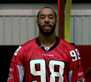 An African American male stands smiling in front of a yellow goal post and net.He is wearing a red and black Atlanta falcons jersey with the number 98 on it in the color white.