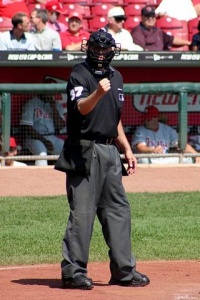 Image Description: An umpire shows the strike symbol dressed in uniform. He stands in front of a baseball dugout. 