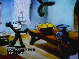 [Image Description: A cartoon scene depicts a cat standing in a room in front of a window and next to a table that holds a bowl and vase. The bowl appears to hold yellow fruits or vegetables. On the bottom end of the screen there are captions that read “Good work, Goldie. Keep it up!” ]