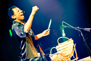 [Image Description: An African American male stands with his mouth open/singing in front of a musical instrument and a microphone on a stand with his fist pumped in the air and drumsticks in his other hand.}