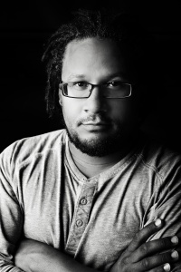 [Image description: A black and white image of an African American man with chin-length dreads, looks into the camera with his arms crossed across his chest. He is wearing dark-rimmed glasses and a three-button shirt.] 