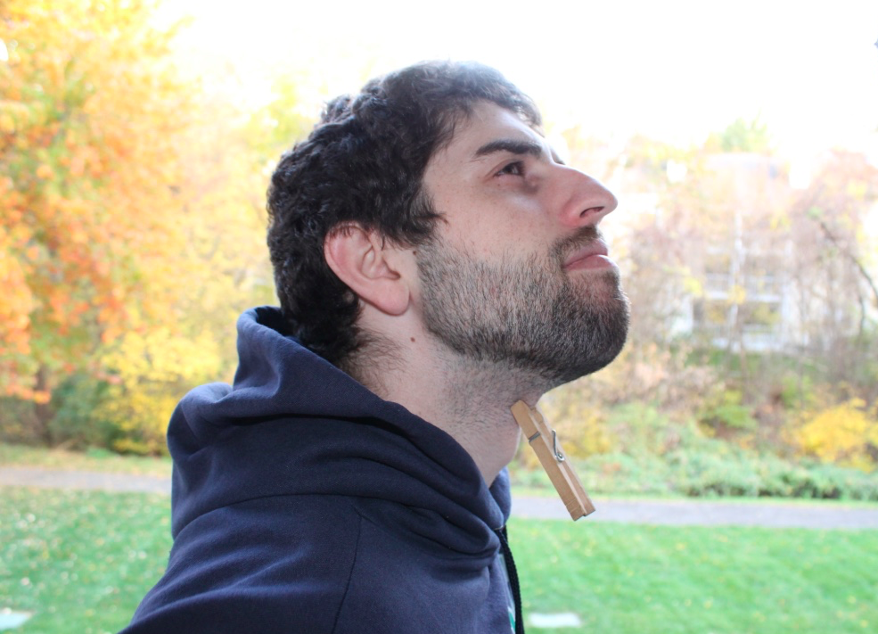 [Image description: A man is standing with their head cocked upward. He is wearing a blue hoodie and on his neck a clothes pin is pinched on his throat. Behind him, trees with fall leaves are visible.]