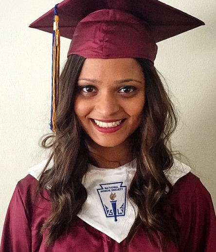 [Image Description: A smiling Indian woman wearing a maroon graduation cap and gown with a white stole looking directly at the camera. Her hair is curled and she is wearing red lipstick.]
