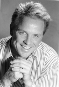 [Image Description: A black and white photograph of a white man with short, light colored hair and light eyes, smiling. His hands are clasped under his chin.]