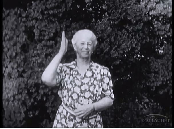 [Image Description: This is a screenshot of a black and white video. A white woman, with grey hair is signing with a small smile on her face.]