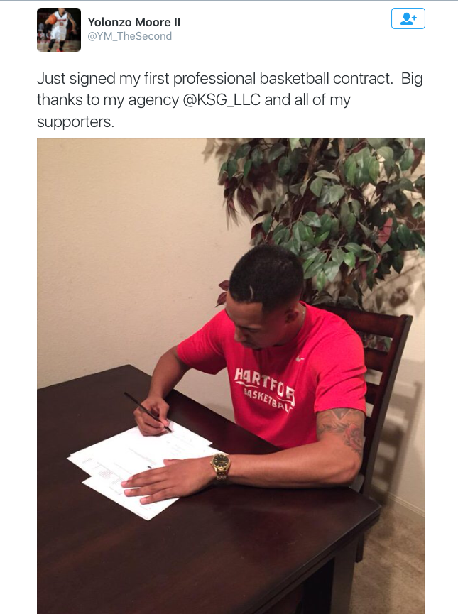 Image Description: A screenshot of a tweet, featuring black text that reads "Just signed my first professional basketball contract. Big thanks to my agency @KSG_LLC and all of my supporters." The tweet includes a large image of a man wearing a red shirt bent over a stack of white papers, holding a pen.]