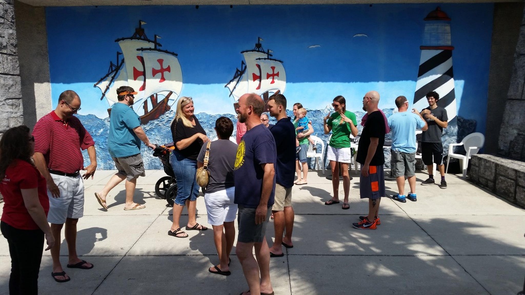 [Image description: A group of adults in shorts and t-shirts gather in the shade, forming conversational circles in front of a large mural. Some parents are holding small children or pushing strollers. On the right, members of the KODAheart team, with green and dark grey t-shirts, can be seen engaging in conversation with groups of adults. In the background, the mural displays 2 pirate ships with red crosses on the white sails on the open water headed toward a black and white striped lighthouse with a red roof.]