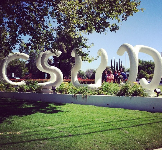 Image description: A brightly lit photo of the "CSUN" sculpture sits on a long white ledge in the grass. Behind it is a group of people, standing between the "U" and "N". On the top left of the photo is a over hanging tree barely touching the beginning of the sculpture.