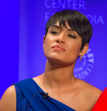 a woman with short hair stands in front of a blue background with a neutral facial expression. Her head is tilted slightly back, and she is wearing lipgloss.]