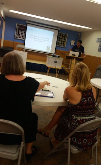 [Image Description: A photo taken behind two people seated at a table facing forward away from the camera. In the background of the photo a man is standing facing the table behind a small podium at a computer. To the left of this is a white backdrop with unreadable projected text.]