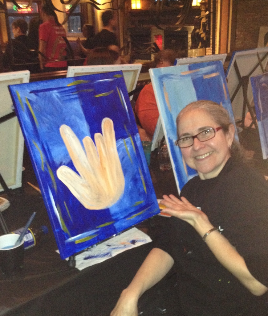 Image description: A women lays her hand flat and gestures towards a painting of an "ILY" hand. the hand is a tan color, while the background is two tones of blue. Behind the hand, the blue background is split vertical and horizontal across the center. The upper left quadrant is a lighter blue, and the right quadrant is a darker blue. The bottom is the reverse of this. Next to the women are a series of other similar paintings.