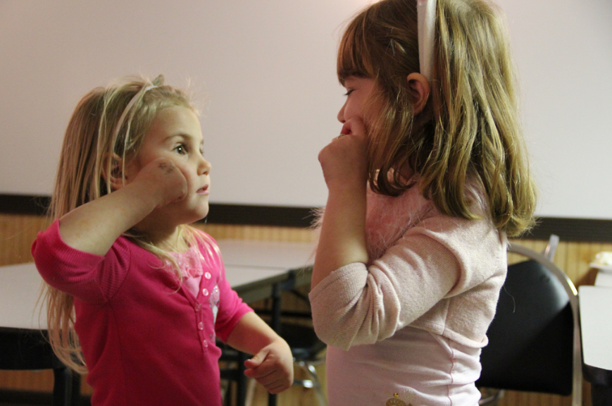 [Image description: A photo of 2 young girls looking at each other while both signing the word apple.There are tables and chairs in the background.]