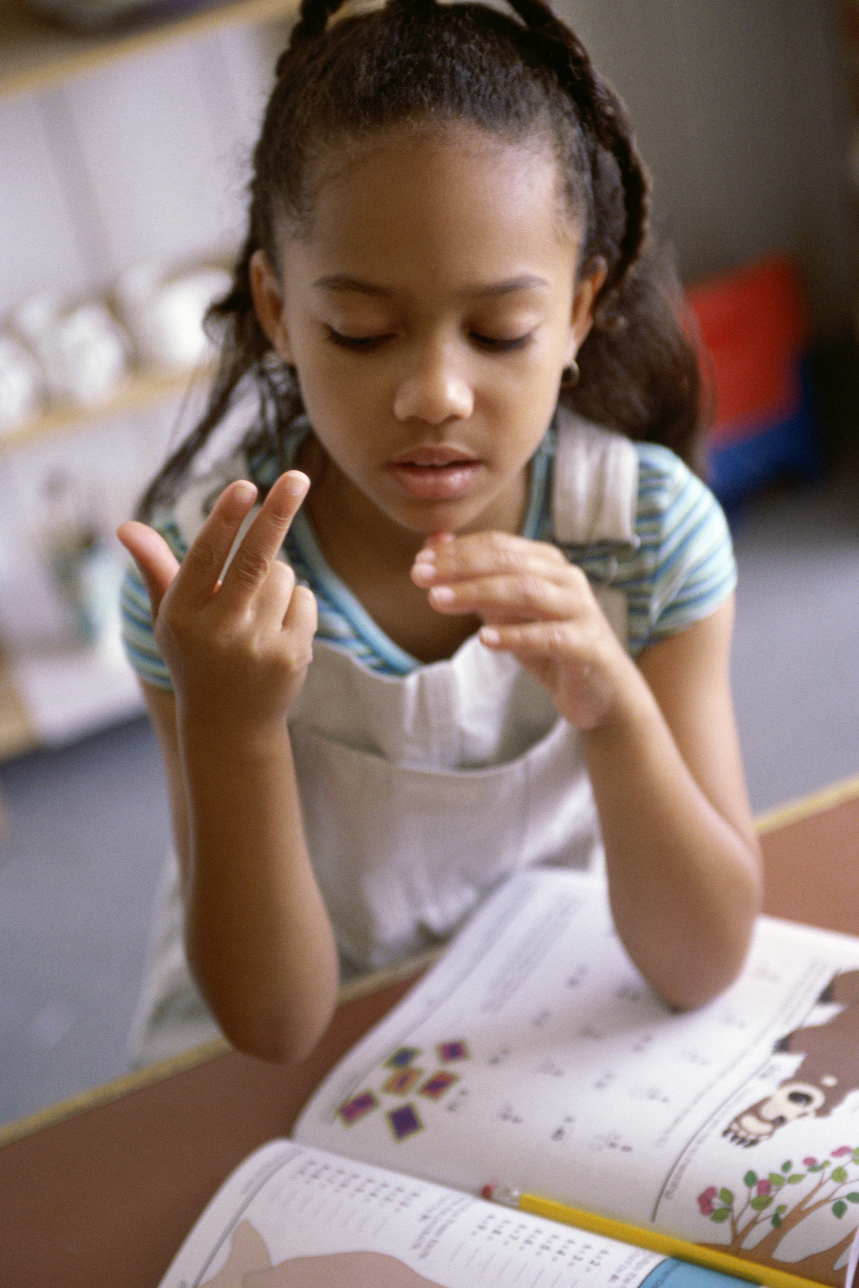 [image description: A young girl is looking down at her hands while forming the sign for 13 with her right hand.The background is blurred a bit.]