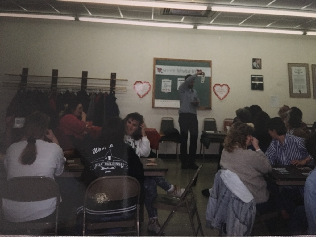 [Image description: In a open room groups of people sit around card tables, at the front of the room a person stands holding a large playing card in the air towards the crowd. There is a small chalkboard in the background.]