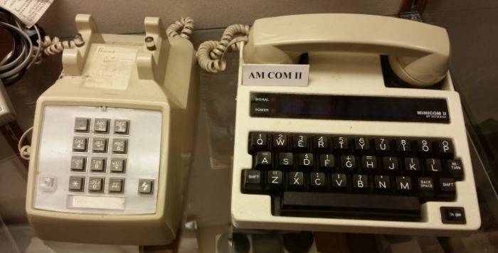 [Image Description: Photo of a beige, landline, touchtone phone with grey buttons, next to a white tty with black keys. The phone’s handset placed on top of the TTY. On top of the TTY is a small white label with black letters reading AM COM II.] 