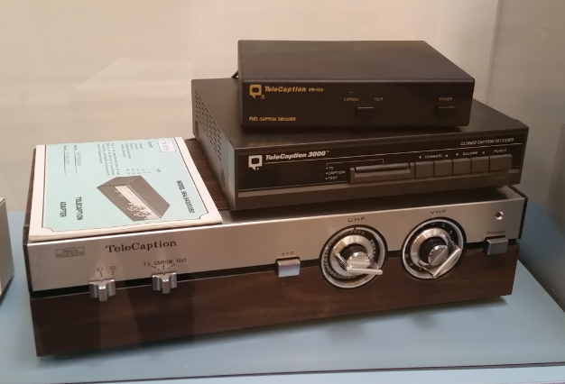 [Image description: Three different versions of closed captioning devices are stacked one on top of another. The older, bigger model is on bottom with two large knobs and buttons. The two smaller, models are encased in black plastic sit on top.]