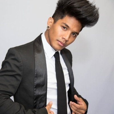 Image Description: A hawaiian male wearing a tuxedo and long black tie looks at the camera head. The angle is tilted to the right and he has a serious look on his face. Both of his hands are opening up his jacket.