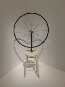 [Image description: a photograph of an art piece featuring a wooden stool with a large wheel on top.]