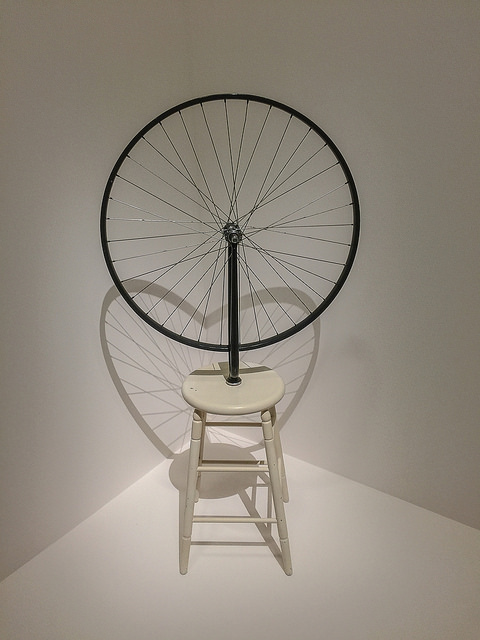 [Image description: a photograph of an art piece featuring a wooden stool with a large wheel on top.]