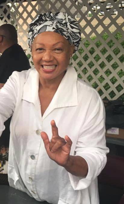 Image Description: An African-American woman wearing a white shirt and a black and white headdress stands smiling and producing the ASL sign for “I love you” with her left hand.