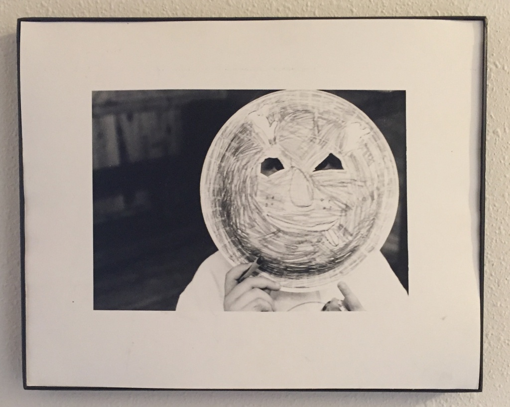 [Image description: an image of a framed black and white photograph. In the photo a child holds a paper plate mask in front of their face.]