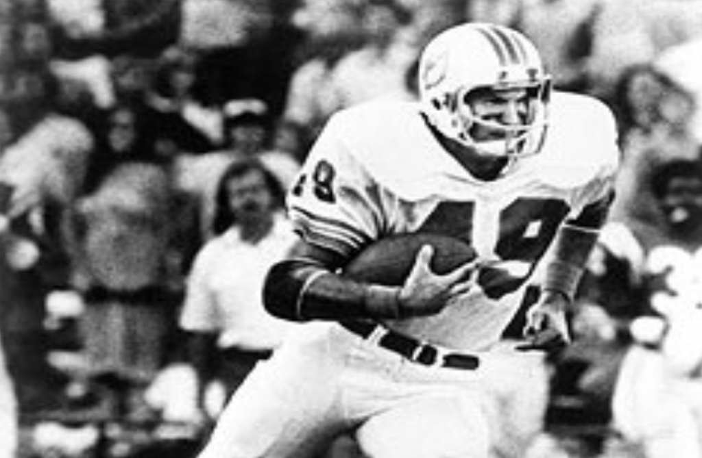 [Image description: Image Description: A black and white photo of a football player in full gear wearing jersey 49 running with the football cradled in his left arm. In the background many fans sitting in the stands watch  in suspense.]