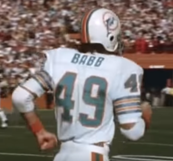 [Image description: Picture of the back of the torso of a fully geared up football player jogging away from the camera on a football field. The back of the white jersey has the last name “BABB” in teal and the numbers “49” in teal outlined in orange. In the background is a blurred rendering of fans in the stadium.] 