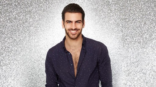 A white man smiling in an unbuttoned navy blue shirt stands facing the camera in front of a sparkly silver background.