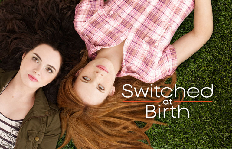 Two white females lay on grass in opposite directions with their heads next to each other. White text to the right of their heads reads“Switched at Birth”. 