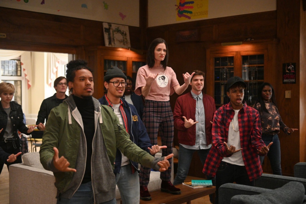 A close cropped photo that shows a group of people standing in a room signing “Now”.