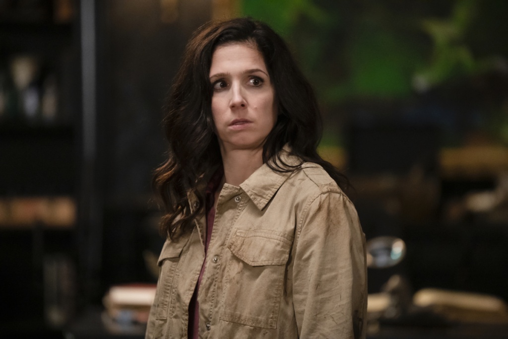 A white woman stands in a tan jacket looking concerned.