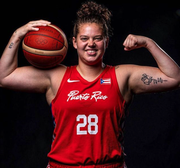 Image Description: A female basketball player with curly hair with both of her arms in an overhead flexing position with her hair in a bun smiles facing the camera . A basketball is between her right bicep and her right hand. On her jersey is “Puerto Rico 28” with the Nike symbol on the left side and Puerto Rican flag on the right side.