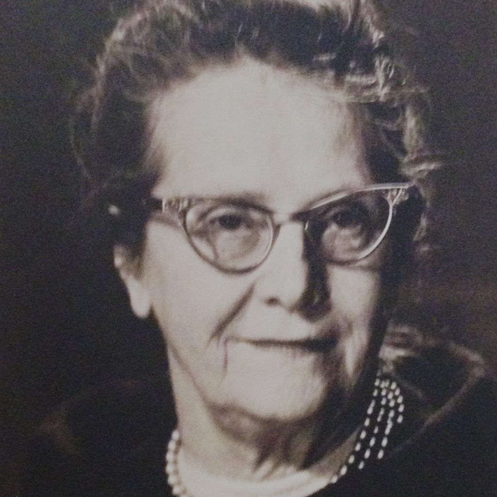 This black and white portrait of Elizabeth Benson shows a white woman with 1960's-style-horn-rimmed glasses and pearl necklace smiling at the camera. Her dark hair is pulled back atop her head and a broad collar encircles her shoulders.
