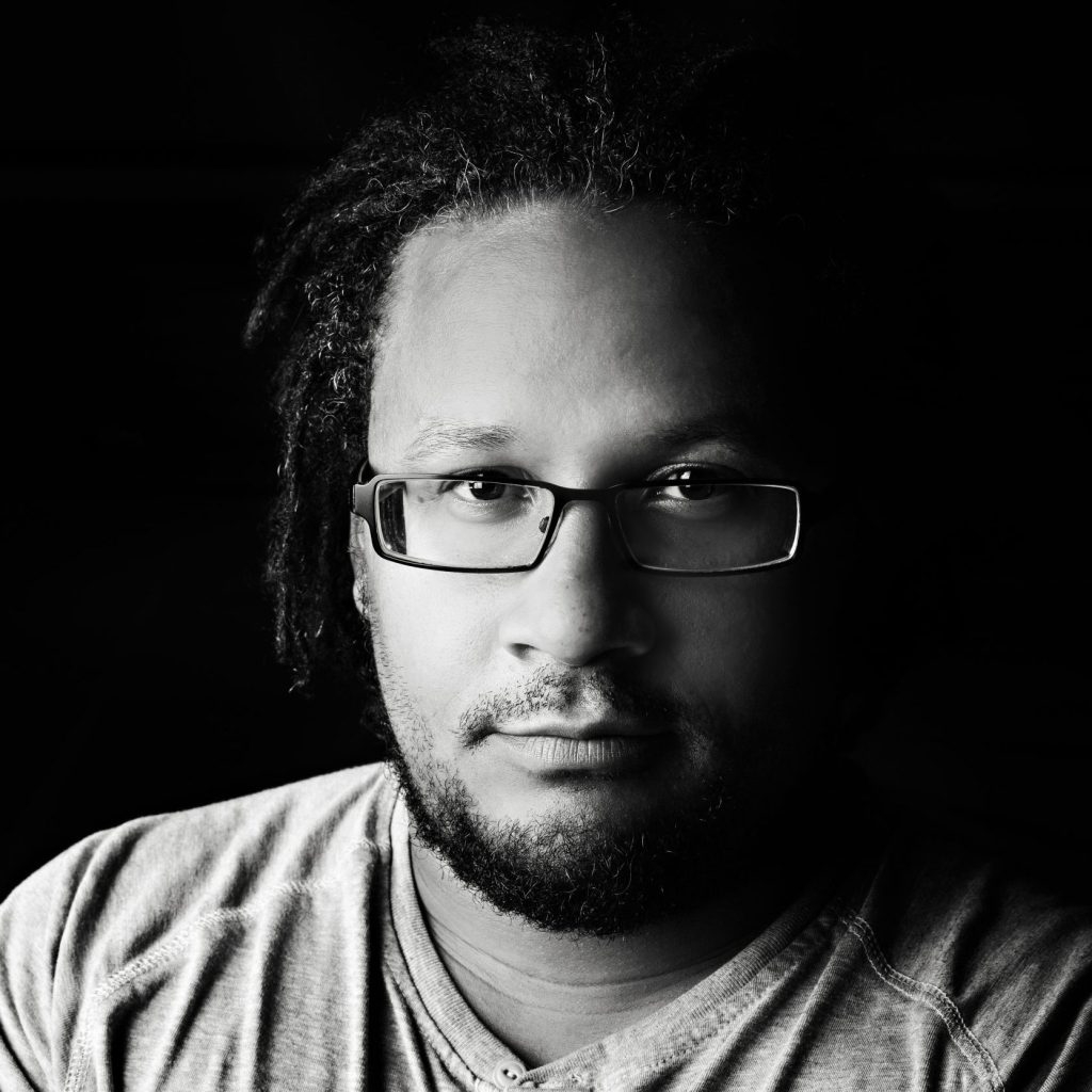 Image description: A black and white image of an African American man with chin-length dreads, looks into the camera with his arms crossed across his chest. He is wearing dark-rimmed glasses and a three-button shirt.
