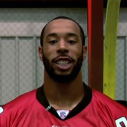 Image description: An African American male stands smiling in front of a yellow goal post and net.He is wearing a red and black Atlanta falcons jersey with the number 98 on it in the color white.