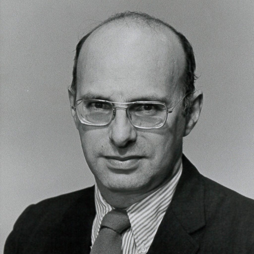Image Description: A black and white photo of an older white male with glasses. He is wearing a suit and tie.