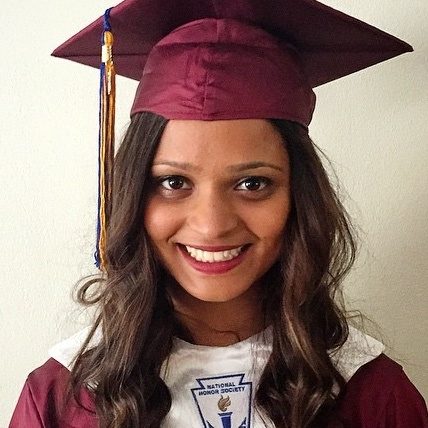 A smiling Indian woman wearing a maroon graduation cap and gown with a white stole looking directly at the camera. Her hair is curled and she is wearing red lipstick.