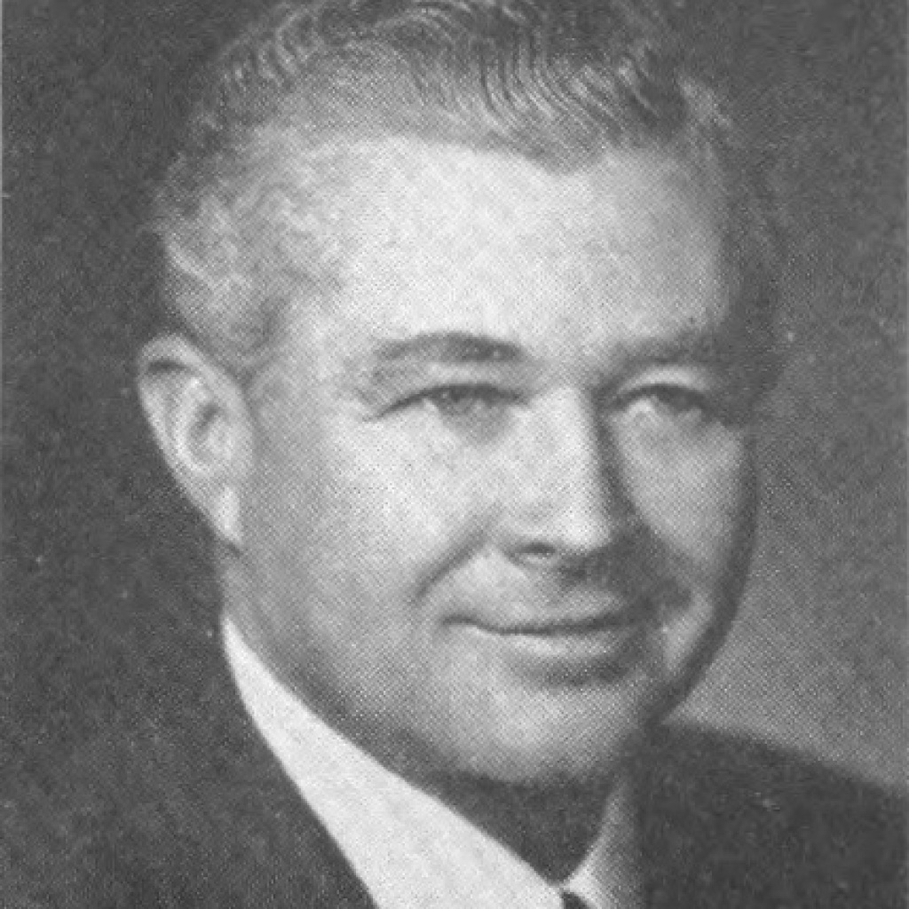 A black and white image of a white man with a suit and tie, smiling at the camera.