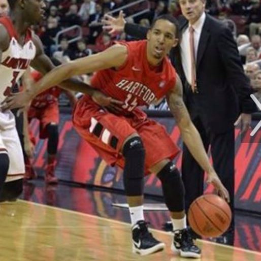 Image description: An action photo of two basketball players on the court. In the foreground, in a red jersey, an African American man dribbles the ball.
